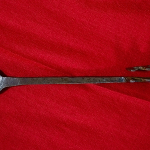Könnte beinhalten: Ein handgeschmiedeter Metalllöffel mit einer abgerundeten Schale und einem hakenförmigen Griff. Der Löffel ist aus dunklem, gealtertem Metall, möglicherweise Eisen oder Stahl, mit rustikalem Aussehen. Der Hintergrund ist ein roter Stoff.