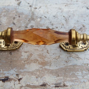 Op de afbeelding: Een antieke kastgreep met een facetgeslepen amberkleurig glazen middenstuk en messing beslag. De messing basisplaten hebben een sierlijk, vintage ontwerp. Deze decoratieve greep is geschikt voor meubelrestauratie of voor een vleugje elegantie.