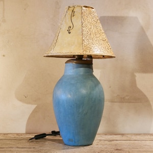 Może przedstawiać: Niebieska ceramiczna lampa stołowa z kremowym abażurem. Abażur ma dekoracyjny wzór i falisty brzeg. Lampa ma podstawę w kolorze mosiądzu i czarny przewód zasilający.