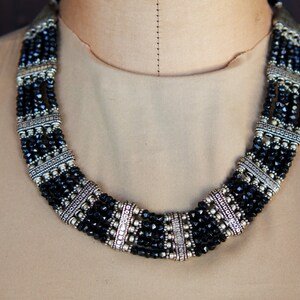 May include: A multi-strand necklace with black beads and silver accents. The necklace is made of several strands of black beads, each separated by a silver spacer. The spacers are rectangular and have a decorative design.