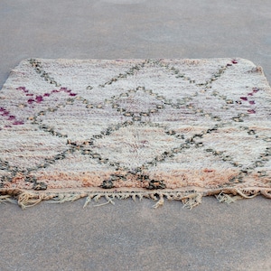 Puede incluir: Una alfombra marroquí beige, blanca y burdeos con un patrón de diamantes. La alfombra tiene un borde de flecos.