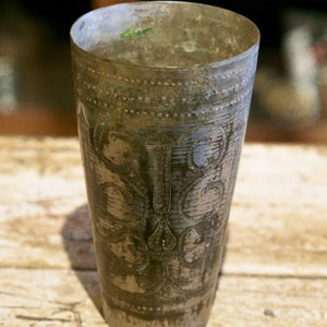 Puede incluir: Un vaso alto de metal plateado antiguo con diseños florales y de volutas grabados. El vaso tiene un aspecto ligeramente deslustrado, lo que sugiere edad y uso. El borde y la base presentan bandas decorativas.