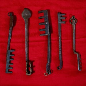 Könnte beinhalten: Sammlung antiker Metallschlüssel in verschiedenen Formen und Größen, angeordnet auf rotem Hintergrund. Die Schlüssel weisen aufwendige Designs auf und weisen Alterserscheinungen auf, was darauf hindeutet, dass es sich um Vintage- oder historische Artikel handelt.