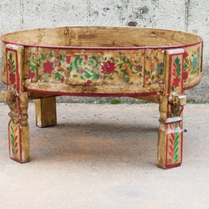Puede incluir: Una mesa redonda de madera con un diseño floral pintado en rojo, verde y amarillo. La mesa tiene cuatro patas y está hecha de madera.