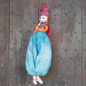 Puede incluir: Un títere azul y naranja con una cara pintada y un tocado rojo y blanco colgando de una puerta de madera.