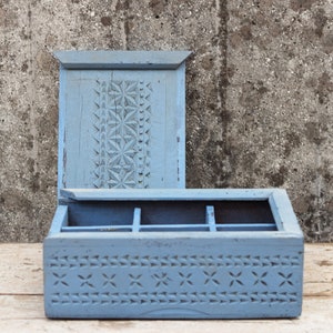 Può includere: Una scatola di legno azzurro con coperchio aperto. La scatola presenta motivi geometrici intagliati sul coperchio e sul davanti. L'interno è diviso in scomparti. La scatola è posta su uno sfondo grigio strutturato.