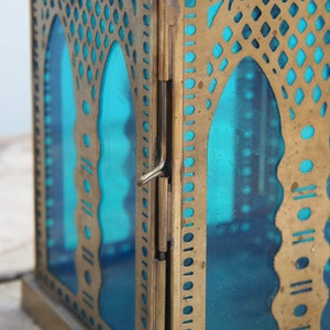 May include: A brass lantern with a teal blue glass panel and intricate cut-out designs. The lantern has a hinged door and a decorative brass frame.