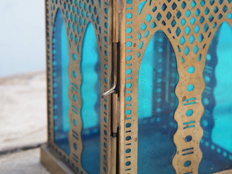 May include: A brass lantern with a teal blue glass panel and intricate cut-out designs. The lantern has a hinged door and a decorative brass frame.