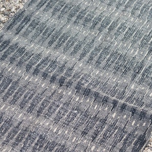 May include: A gray and white patterned rug with a textured woven design. The rug is folded in half and is lying on a bed of small white rocks.