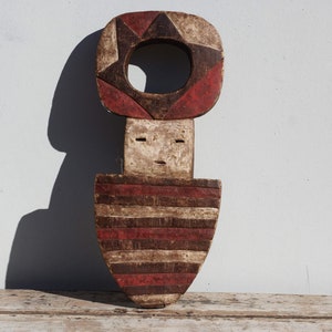 Puede incluir: Una escultura de madera de una figura humana estilizada con una abertura circular en la cabeza. La escultura está pintada en rayas rojas, blancas y negras.
