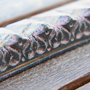 Puede incluir: Primer plano de un marco de madera con un diseño tallado decorativo. La talla presenta un patrón repetitivo de hojas y espirales, pintadas en tonos de rosa y gris.