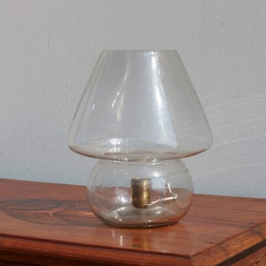 Può includere: Una lampada da tavolo in vetro trasparente a forma di fungo con una base rotonda e un paralume largo e svasato.