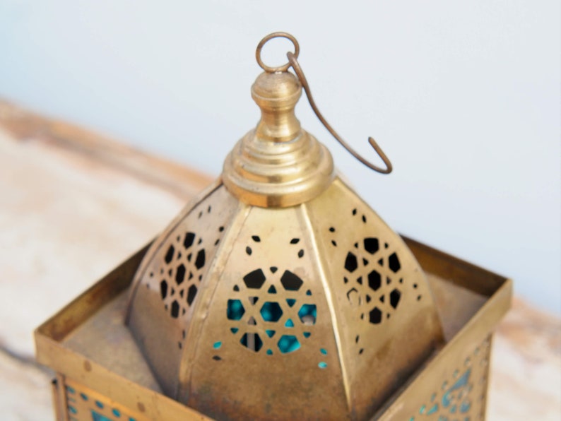May include: A brass lantern with intricate cut-out designs and a turquoise glass panel. The lantern has a hanging loop and a domed top.