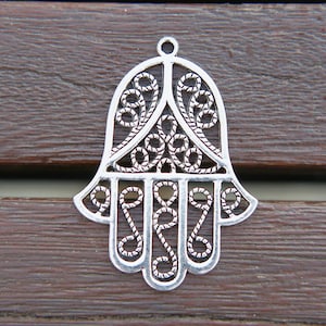 Może przedstawiać: Srebrny metalowy amulet w kształcie ręki hamsa, tradycyjny symbol ochrony ze Środkowego Wschodu. Amulet jest misterny zdobiony wzorem filigranowym.