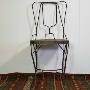 Puede incluir: Una silla de metal vintage con una estructura de alambre oscuro y retorcido y un asiento de madera. La silla tiene una apariencia única y artesanal, con un respaldo alto y elementos decorativos. La silla está colocada contra una pared blanca y una alfombra estampada.