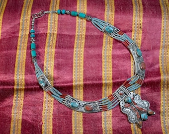 May include: A silver necklace with turquoise and red stones, set against a striped fabric background.