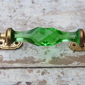 Op de afbeelding: Een decoratieve kastgreep met een facetgeslepen, smaragdgroen glazen middenstuk. De handgreep wordt geflankeerd door messingkleurige metalen houders met decoratieve details. De hardware is ontworpen voor installatie op een kast of lade.