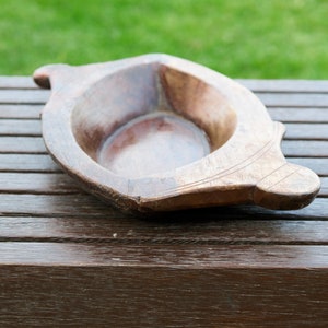 Puede incluir: Un cuenco de servir de madera con forma redondeada y un acabado liso de color marrón oscuro. El cuenco tiene un borde ligeramente elevado y un diseño decorativo tallado en el lateral.