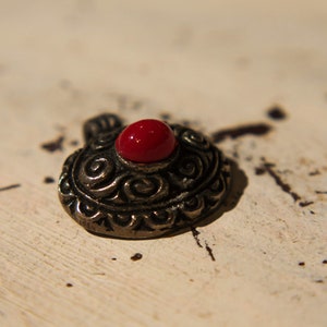 Może przedstawiać: Srebrny charm z misternym wzorem i czerwonym kamieniem szlachetnym w środku.