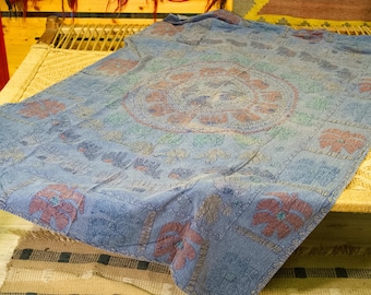 May include: A blue and white patterned throw blanket with a floral design. The blanket is folded and lying on a woven wicker surface.