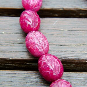 May include: A strand of pink oval-shaped beads with a carved design. The beads are strung on a cord and are lying on a wooden surface.