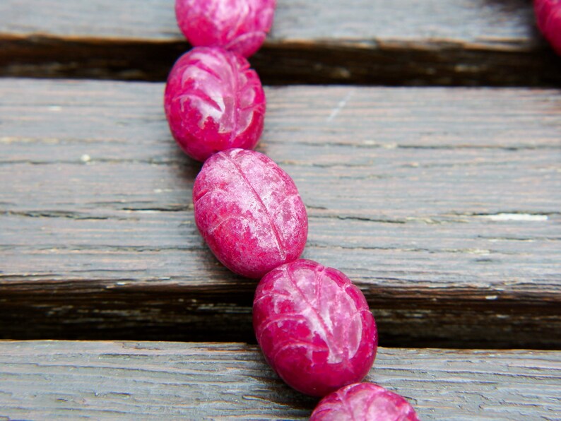 May include: A strand of pink oval-shaped beads with a carved design. The beads are strung on a cord and are lying on a wooden surface.