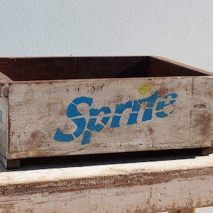 Può includere: Una cassa di legno vintage con la parola "Sprite" dipinta in blu e giallo sul lato. La cassa è usurata e mostra segni di età.