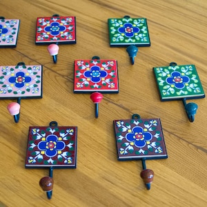 Puede incluir: Un conjunto de ganchos de pared decorativos con azulejos cuadrados de cerámica con diseños florales en varios colores. Cada gancho tiene una base de metal y un pomo decorativo. Los ganchos están colocados sobre una superficie de madera.