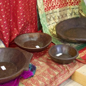 Puede incluir: Cuatro cuencos de madera de diferentes tamaños se exhiben sobre una tela roja y dorada estampada. Los cuencos son de color marrón oscuro y tienen un aspecto rústico tallado a mano. El fondo presenta telas drapeadas de colores en rojo, verde y morado.