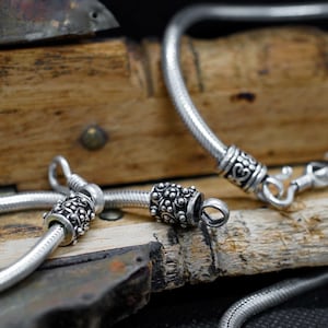 May include: A silver-toned snake chain bracelet featuring intricate, oxidized metal end caps with a detailed floral-inspired pattern. The bracelet includes a secure hook-and-eye clasp mechanism, showcasing a smooth, flexible chain design.