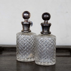 Könnte beinhalten: Zwei klare Glasflaschen mit silbernen Stopfen und einem Diamantenmuster. Die Flaschen stehen auf einer dunklen Holzoberfläche.