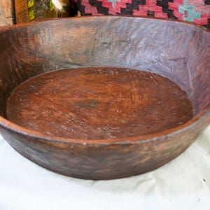 Puede incluir: Un cuenco de madera rústico y grande con acabado marrón oscuro. El cuenco tiene un diseño ancho y poco profundo con lados redondeados y pequeñas asas. La veta de la madera es visible, lo que añade un aspecto natural.