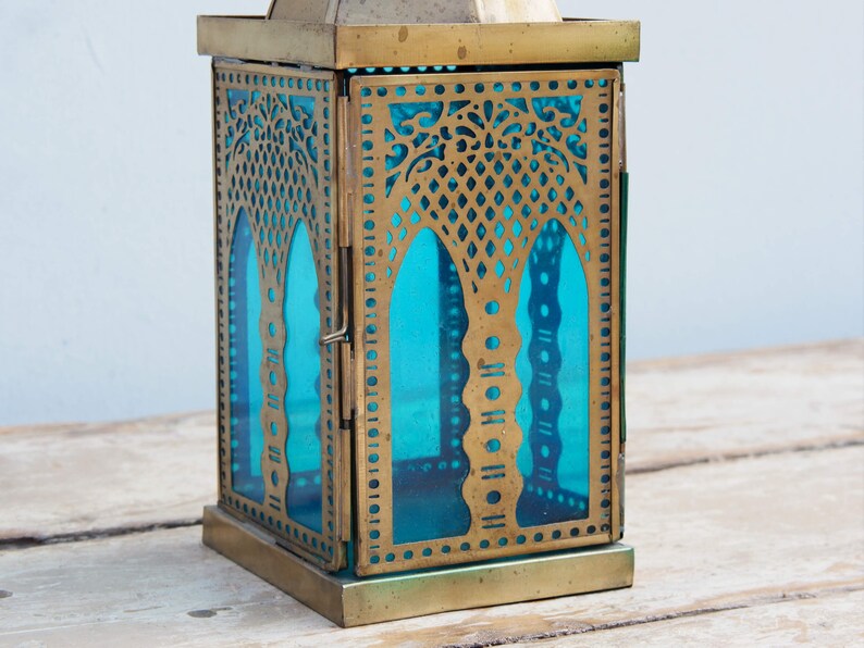 May include: A brass lantern with a blue glass panel and intricate cut-out designs. The lantern has a square base and a hinged door.