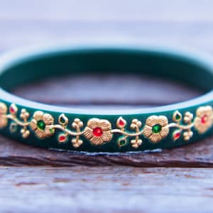 Puede incluir: Un brazalete verde con un diseño floral dorado y rojo. El brazalete está decorado con pequeñas piedras preciosas rojas y verdes.