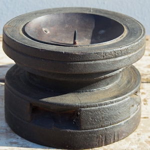 Può includere: Un portacandele in legno scuro con la parte superiore rotonda e un inserto in metallo. Il portacandele ha un aspetto rustico e invecchiato.
