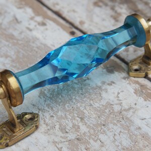 Op de afbeelding: Een decoratieve deurkruk met een facetgeslepen, aquamarijnkleurige glazen handgreep. De handgreep is gemonteerd op messingkleurige metalen beugels. De deurkruk is ontworpen voor meubels of kastdeuren.