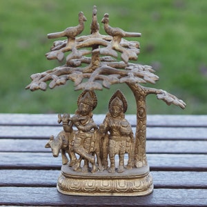 Può includere: Una scultura in metallo dorato di due figure in piedi sotto un albero con uccelli appollaiati sui rami. Le figure tengono strumenti musicali e una mucca è in piedi vicino a loro.