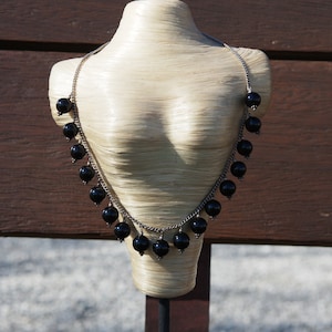 May include: A silver chain necklace with black beads hanging from it. The necklace is displayed on a white mannequin bust.