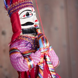 Puede incluir: Un títere tradicional indio con la cara pintada, que lleva un traje estampado en rojo, rosa y azul. El títere se está llevando el dedo a los labios, como para decir "shhh".