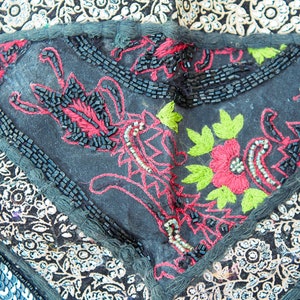 May include: Close-up of a black fabric with intricate embroidery in red, green, and white thread. The fabric is decorated with black beads and sequins.