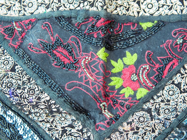 May include: Close-up of a black fabric with intricate embroidery in red, green, and white thread. The fabric is decorated with black beads and sequins.