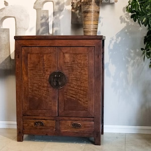 May include: A dark brown wooden cabinet with two doors and two drawers. The cabinet has a circular metal handle and decorative hardware. A vase with dried flowers sits on top. The cabinet is against a light gray wall.
