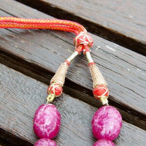 May include: Close-up of a pink gemstone necklace with gold and red accents. The necklace has a tassel-like closure with a red and gold cord.