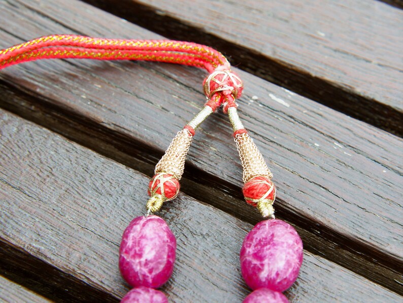 May include: Close-up of a pink gemstone necklace with gold and red accents. The necklace has a tassel-like closure with a red and gold cord.