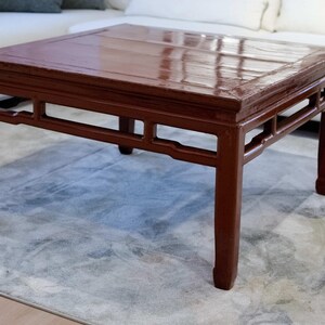 Puede incluir: Una mesa de centro cuadrada de madera de color marrón rojizo oscuro. La mesa tiene un acabado brillante y una parte superior rectangular. Las patas son rectas y robustas, con recortes decorativos. La mesa mide aproximadamente 76 cm cuadrados y 46 cm de alto.