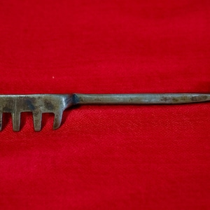 Könnte beinhalten: Ein antikes Metallwerkzeug mit kammähnlichem Ende und Schlaufengriff. Das Werkzeug hat einen langen, schlanken Griff und eine Reihe von vier gleichmäßig verteilten Zinken. Das Metall hat ein angelaufenes Aussehen, was auf Alter hindeutet. Das Werkzeug ist vor rotem Hintergrund platziert.