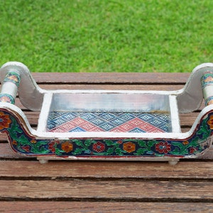 Puede incluir: Una bandeja plateada con diseños florales pintados en tonos de azul, verde, rojo y amarillo. La bandeja tiene dos asas y un inserto de vidrio transparente.
