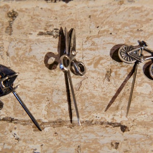 Może przedstawiać: Trzy srebrne szpilki z różnymi wzorami: żaglówka, para nożyczek i rower. Szpilki znajdują się na jasnobrązowej drewnianej powierzchni.