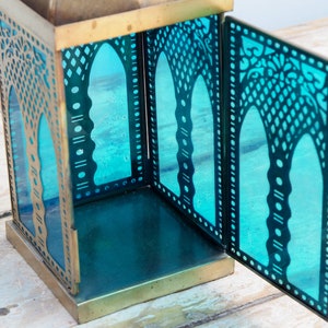 May include: A brass lantern with a teal blue glass insert. The lantern has intricate cutouts in the metal and glass, creating a decorative pattern. The lantern is open, revealing the empty interior.
