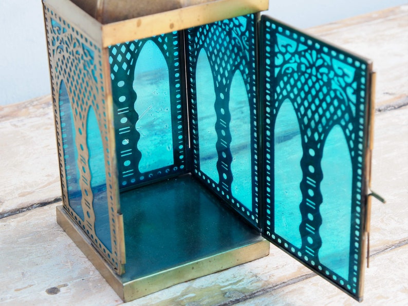 May include: A brass lantern with a teal blue glass insert. The lantern has intricate cutouts in the metal and glass, creating a decorative pattern. The lantern is open, revealing the empty interior.